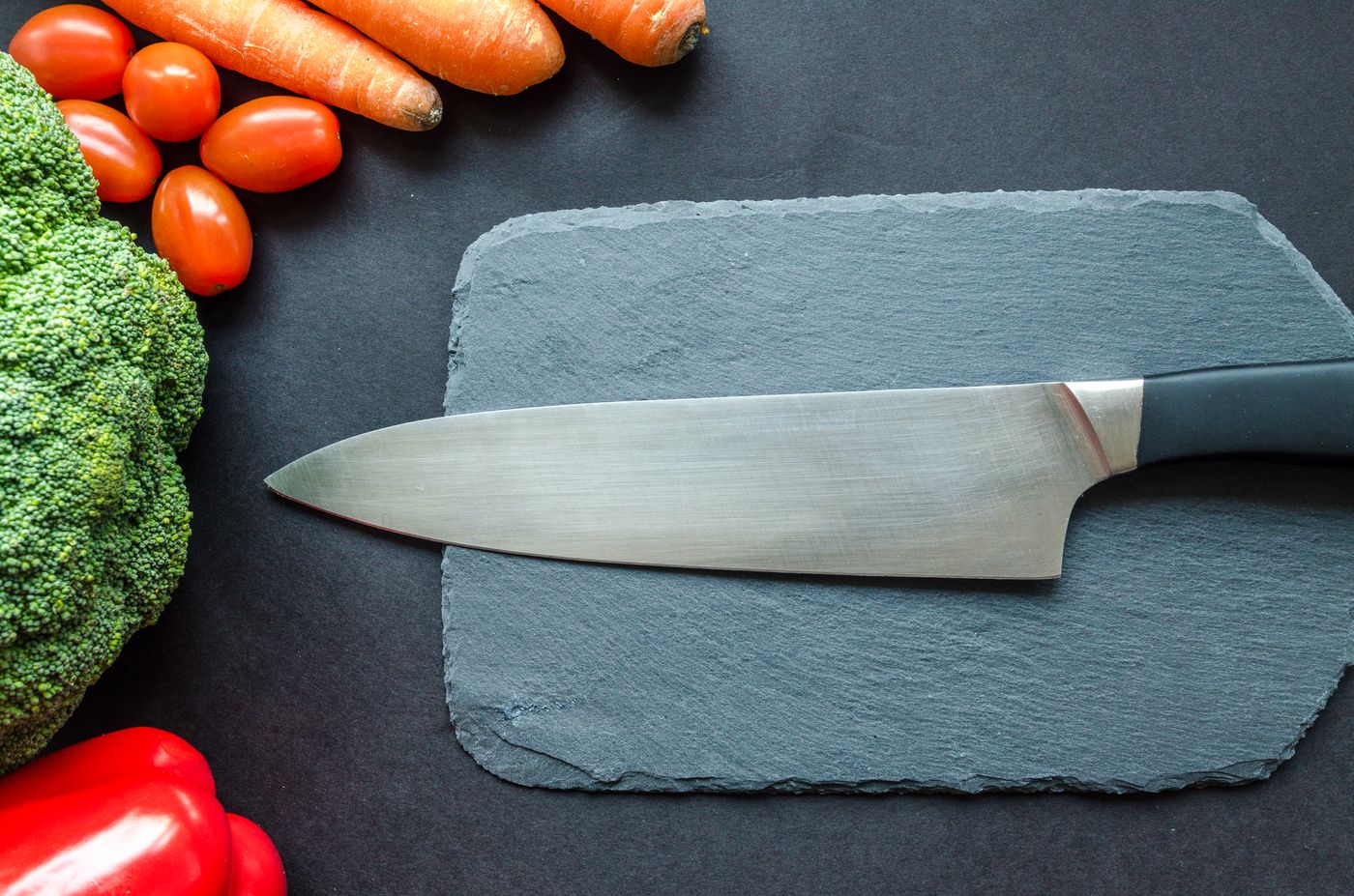 Actual chef knife beside vegetables on a slate board.